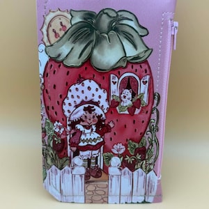 May include: Pink wallet featuring a strawberry house design with a cartoon character, a white picket fence, and a cat in a window. The wallet has a zipper closure and a small tab closure.