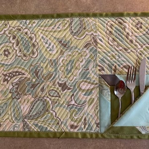 Op de afbeelding: Een groene en bruine placemat met paisley-patroon met een groene stoffen zak aan de rechterkant. De zak heeft een bruine rand en bevat een zilveren vork, mes en lepel.