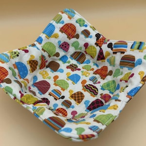 May include: A square fabric bowl with a white background and a colorful turtle pattern. The turtles are in various colors, including blue, green, orange, and brown. The bowl is designed to hold items.