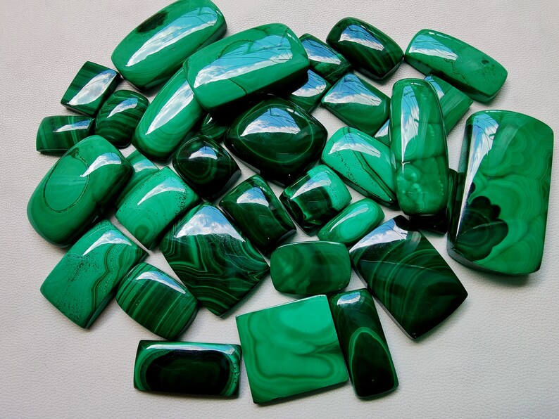 May include: A collection of 20 green malachite cabochons, each with a unique pattern of veins and swirls. The stones are smooth and polished, with a glossy finish. They are ideal for jewellery making or other craft projects.