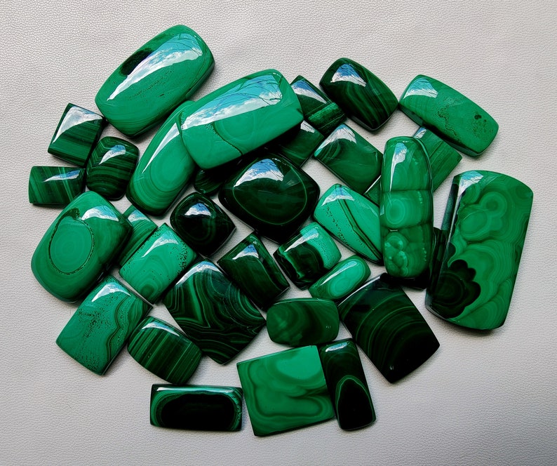 May include: A collection of 20 green malachite cabochons, each with unique patterns and veining. The stones are various shapes and sizes, ranging from small to large.