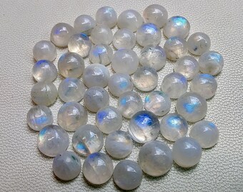 Puede incluir: Una colección de cabochones redondos de piedra lunar blanca con destellos iridiscentes azules y verdes. Las piedras son lisas y pulidas, y están dispuestas en una pila suelta.