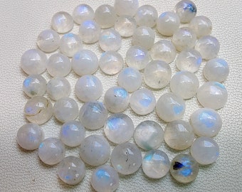 Puede incluir: Una colección de cabujones redondos de piedra lunar blanca con destellos iridiscentes azules y blancos. Las piedras están dispuestas en círculo sobre una superficie blanca.