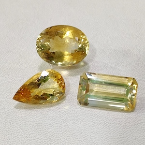 Könnte beinhalten: Drei facettierte Edelsteine in Gelb- und Goldtönen. Die Formen umfassen ein Oval, eine Birne und einen rechteckigen Smaragdschliff. Die Edelsteine werden auf einer weißen Oberfläche präsentiert.