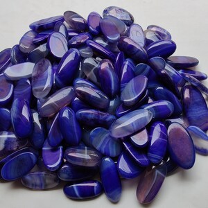 May include: A pile of polished, oval-shaped gemstones in shades of deep blue and purple. The stones have a glossy finish and display natural banding patterns, creating a marbled effect. The gemstones are likely used for jewelry or decorative purposes.