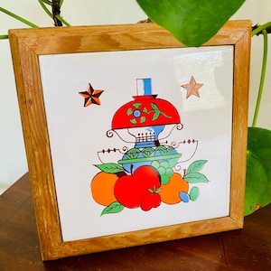 May include: Square, framed tile featuring a vibrant illustration of a red-topped lamp, fruit, and stars. The tile is set in a light brown wooden frame, suitable for kitchen or dining room decor.