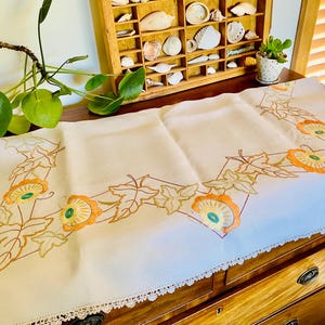 May include: Cream-coloured embroidered table runner featuring an orange and yellow floral design with green leaves. The runner has a delicate lace trim. The embroidery is detailed, adding a vintage touch to the home decor.