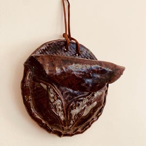 May include: A circular, dark brown glazed ceramic wall pocket with a glossy finish. The piece features a folded upper section and a lower area with white, textured, organic-looking details. A thin brown leather cord is attached for hanging.