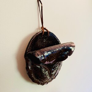May include: A dark brown ceramic wall pocket with a textured surface and a curved opening. It is suspended by a brown leather cord. The interior has a glossy, iridescent finish.