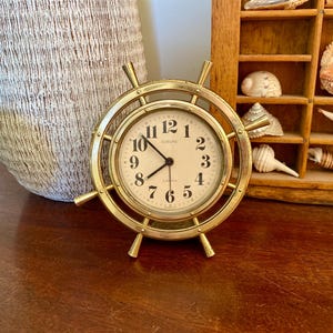 May include: A gold-coloured clock shaped like a ship's wheel. The clock face is cream with black numerals and hands. The clock sits on a wooden surface, with a decorative vase and a shelf of seashells in the background.