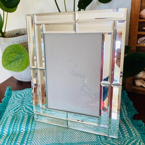 May include: A rectangular mirrored picture frame with a beveled edge design. The frame is made of glass and has a reflective surface. The frame is on a turquoise and white patterned placemat.