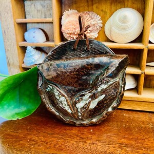 May include: A brown ceramic wall pocket with a textured surface and decorative details. It hangs from a brown leather cord. The background features a wooden shelf with seashells.