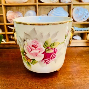 May include: A white porcelain planter with a gold rim and handles, featuring a hand-painted design of pink roses and green leaves. The planter has a rounded shape and sits on small feet. Decorative item.