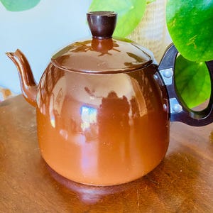 May include: A brown vintage teapot with a rounded body, spout, and handle. The lid has a dark brown knob. The teapot is made of a shiny material and sits on a wooden surface. The handle is black and attached with silver rivets.