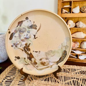 May include: A large, round ceramic plate with a beige background and abstract brown, white, and green painted design. The plate is displayed on a wooden stand. Decorative item for the home.