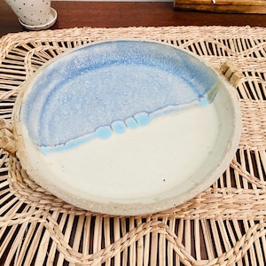 May include: Handmade ceramic serving dish with a two-tone design. The top half is light blue, transitioning to white. Features two small handles and a textured rim. Placed on a woven placemat.