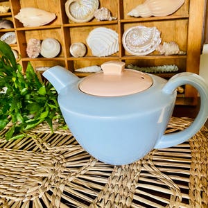 May include: A light blue teapot with a pink lid. The teapot is sitting on a woven straw mat.
