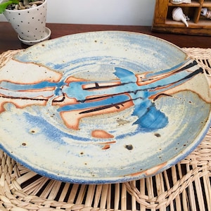 May include: A handcrafted ceramic plate with an abstract design. The plate has a cream base with blue, orange, and black brushstrokes. The rim is a darker blue. This decorative plate is suitable for serving food or as a display item.