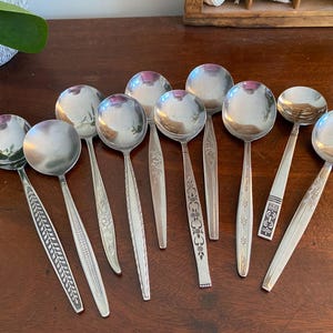 May include: A collection of nine silver-toned metal spoons arranged in a fan shape. Each spoon features a unique embossed or engraved pattern on the handle, ranging from floral motifs and leaf designs to geometric lines and textured borders.