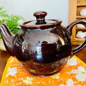 May include: A glossy dark brown ceramic teapot with a matching lid, featuring a rounded body, a curved spout, and a sturdy handle. The teapot has a smooth, reflective surface and is positioned on an orange fabric mat with a white floral pattern.