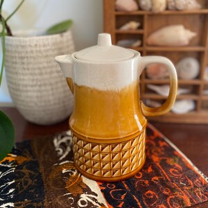 May include: A vintage ceramic coffee pot with a cream lid and handle, and a two-tone body in cream and amber. The lower section has a geometric pattern. The pot rests on patterned fabric, with a plant and a decorative shelf in the background.