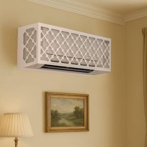Op de afbeelding: Witte decoratieve airconditioner cover met een geometrisch traliewerkontwerp, gemonteerd op een beige muur. De cover verbergt de airconditioning unit. Een ingelijst landschapsschilderij en een lamp zijn ook zichtbaar.