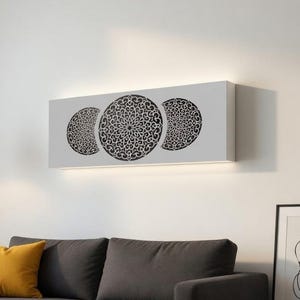 May include: A contemporary living room setting with a grey sofa and a yellow cushion. Above the sofa, a rectangular wall art piece showcases three circular, black, ornate designs on a white backdrop. A black floor lamp and framed abstract art complete the decor.