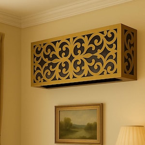May include: A gold-coloured rectangular decorative cover featuring a black, ornate scrollwork design, mounted on a beige wall. A framed landscape painting and a lamp with a beige shade are below the cover.