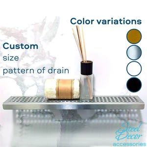 May include: A stainless steel shower shelf with a grid pattern. The shelf is mounted on a white tiled wall. The shelf is empty except for a white towel and a glass bottle with a black lid. The text "Custom size pattern of drain" is visible on the left side of the image. The text "Color variations" is visible on the right side of the image. Four colour swatches are visible: gold, silver, white, and black.