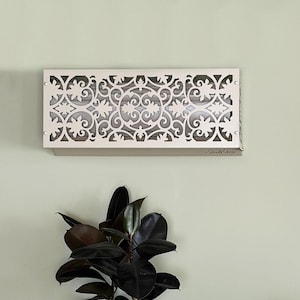 May include: Cream-coloured, rectangular wall sconce with an intricate floral and scroll cut-out design. The sconce is mounted on a light green wall. The bottom right corner of the sconce has the text "Brookhaven".