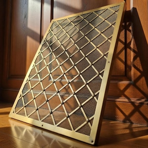 Può includere: Uno schermo metallico dorato rettangolare con un motivo a rombi. Lo schermo è appoggiato a una porta e un pavimento in legno, con la luce del sole che proietta un'ombra. La cornice è di un caldo colore oro e la rete è un bronzo più scuro.