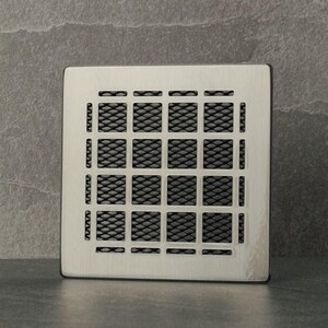 Op de afbeelding: Een vierkante zilveren ventilatiekap met een zwart metalen gaas.