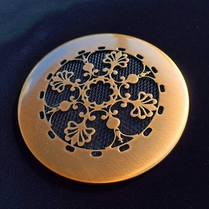 Könnte beinhalten: Ein rundes, bronzefarbenes dekoratives Metallstück mit einem aufwendigen floralen Design. Die Mitte hat ein dunkles Netz, und der äußere Rand hat kleine, gleichmäßig verteilte Aussparungen. Es ist wahrscheinlich ein dekoratives Element oder eine Abdeckung.