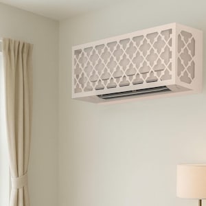 May include: A white air conditioning unit cover with a geometric design is mounted on a light-coloured wall. The cover has a rectangular shape and a lattice-like pattern. A window with beige curtains is visible to the left, and a lamp is on the right.