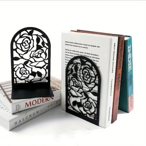 Peut inclure: Des serre-livres en métal noir avec un motif de roses, soutenant des livres aux dos blancs et colorés. Les livres sont intitulés "MODERN" et "HALCNOWBC ODEAR".