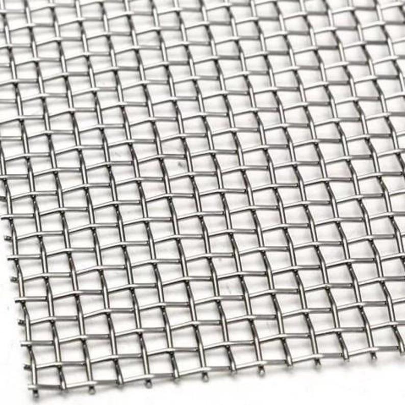 May include: Close-up of a stainless steel mesh screen. The screen is made of interwoven metal wires, forming a grid pattern. The mesh has a square pattern and a metallic silver color.