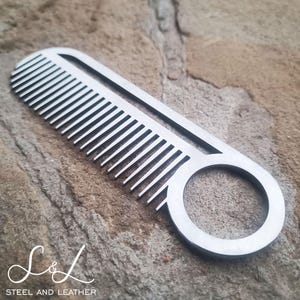 May include: A silver metal comb with a round hole in the handle. The comb is on a rough, brown surface. "Steel and Leather" is written in a cursive font below the comb.
