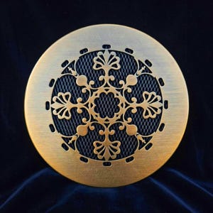 Peut inclure: Un couvercle de ventilation rond en métal de couleur laiton antique avec un motif floral complexe. Le centre présente un maillage sombre et le bord extérieur comporte de petits trous espacés. Le design est symétrique et détaillé.