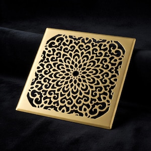 May include: A square, gold-coloured metal decorative grate featuring an elaborate, symmetrical floral design. The openwork pattern reveals a dark background, creating a striking contrast. The grate has a smooth, polished finish and a slightly raised edge.