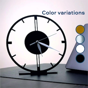 May include: A black metal clock with white hands and a black center circle. The clock is on a stand and has a white background.