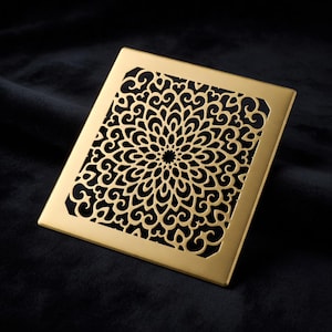 May include: A square, gold-coloured metal decorative grate with an intricate floral pattern. The design features a central circular motif surrounded by swirling, symmetrical shapes. The grate is set against a dark, textured background.