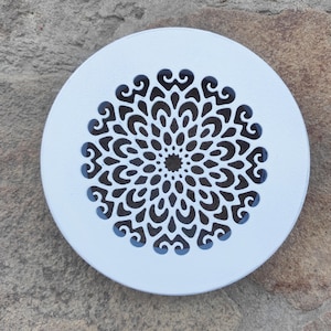 Puede incluir: Una cubierta de ventilación decorativa blanca y circular con un patrón floral simétrico detallado recortado en el centro. El diseño presenta una serie de formas curvas y una abertura central circular. La cubierta es de metal.