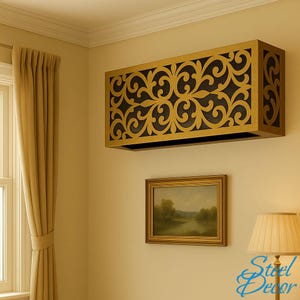 Op de afbeelding: Een rechthoekige, goudkleurige metalen airconditioner cover met een zwart, sierlijk, krulwerk ontwerp. De cover is gemonteerd op een beige muur boven een ingelijste landschapsschilderij. Een beige gordijn en een lamp met een beige lampenkap zijn ook zichtbaar.
