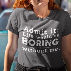 May include: A dark gray t-shirt with the text "Admit it: Life would be BORING without me." The shirt is worn by a person with red hair and sunglasses, leaning on a teal car.
