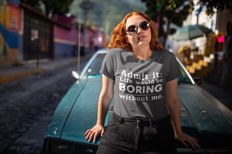 May include: A dark gray t-shirt with the text "Admit it: Life would be BORING without me." The shirt is worn by a person with red hair and sunglasses, leaning on a teal car.