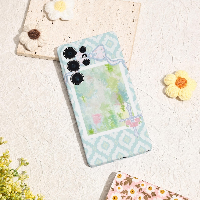May include: A light blue phone case with a white geometric pattern and a watercolor design. The case features a bow and floral accents. The phone case has a camera module with multiple lenses.