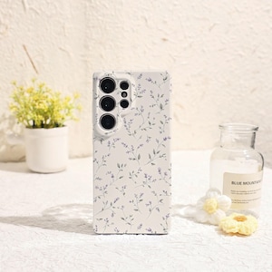 Peut inclure: Coque de téléphone blanche ornée d'un motif floral de petites fleurs violettes et de feuilles vertes. L'étui comporte des découpes pour les objectifs de l'appareil photo et les boutons du téléphone. Design floral.