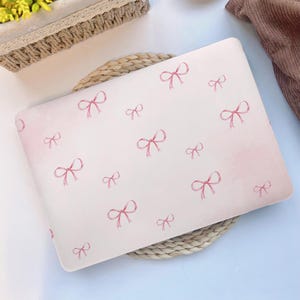 Könnte beinhalten: Eine weiße Laptop-Hülle mit einem Muster aus rosa Aquarell-Schleifen. Die Schleifen sind über die Hülle verteilt, mit einem weichen rosa Farbverlauf im Hintergrund. Die Hülle liegt auf einer gewebten Matte.