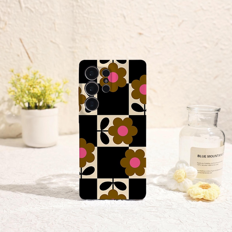 May include: A phone case with a retro floral pattern. The design features brown flowers with pink centers set against a black and cream checkered background. The case has a camera module with three lenses.