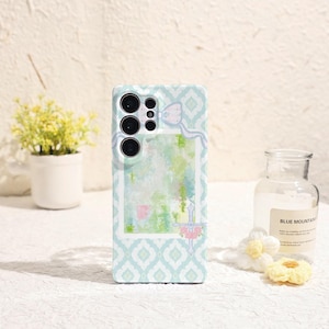 May include: A light blue phone case with a white and blue geometric pattern. The case features a decorative frame with a watercolor design in shades of green, blue, and pink. A bow and floral accents complete the design.